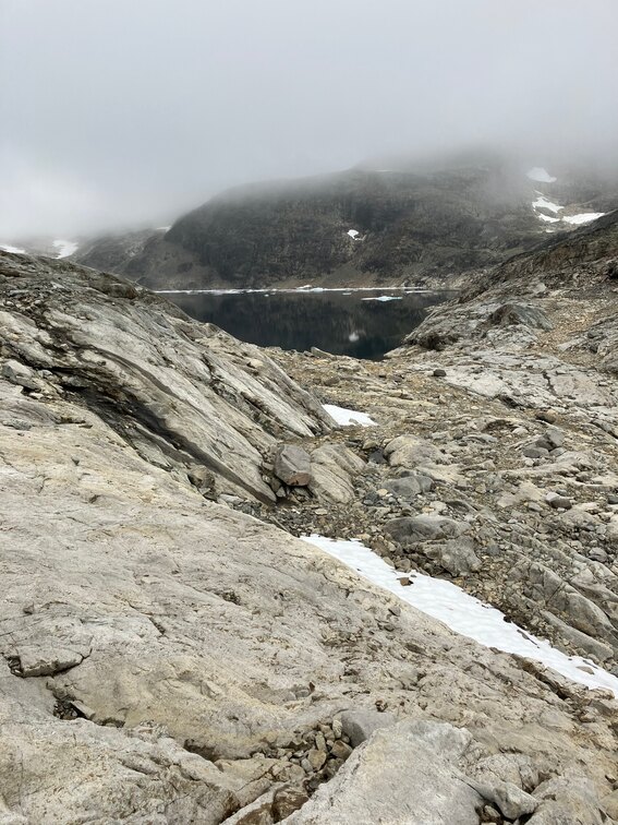 Stony alpine landscape with small snowfields, a still dark lake in the background with floating ice. Low clouds